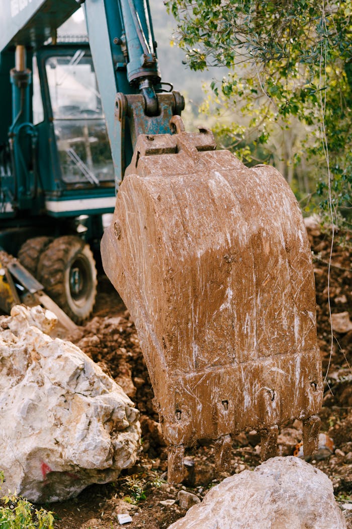 about-us-01 Close-up of a muddy excavator bucket at a construction site, surrounded by rocks and earth.